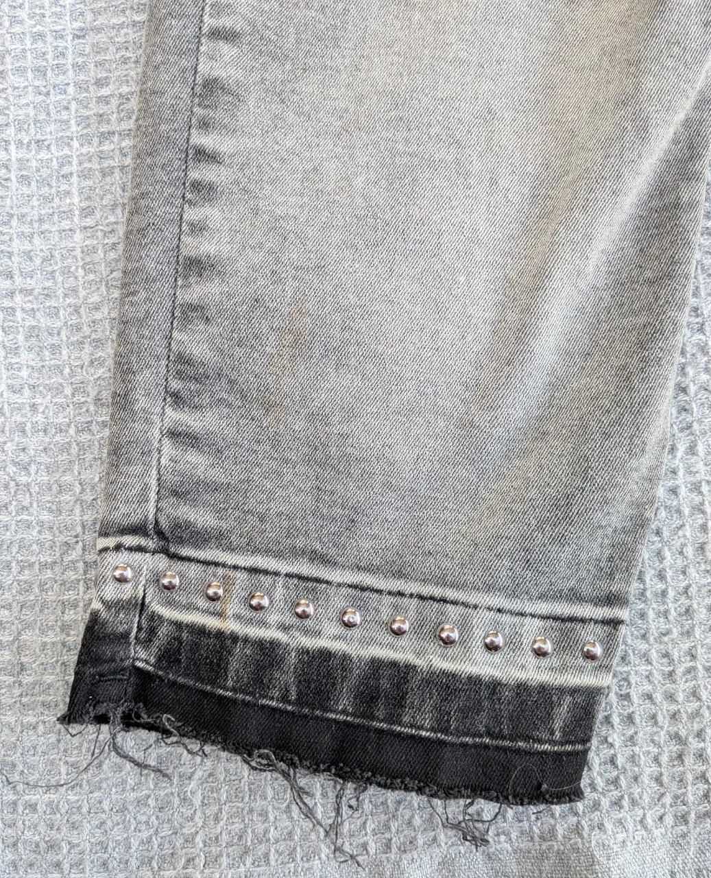 Jeans grises con tachas plateadas en los bajos y terminación deshilachada. Le falta el cursor al cierre.