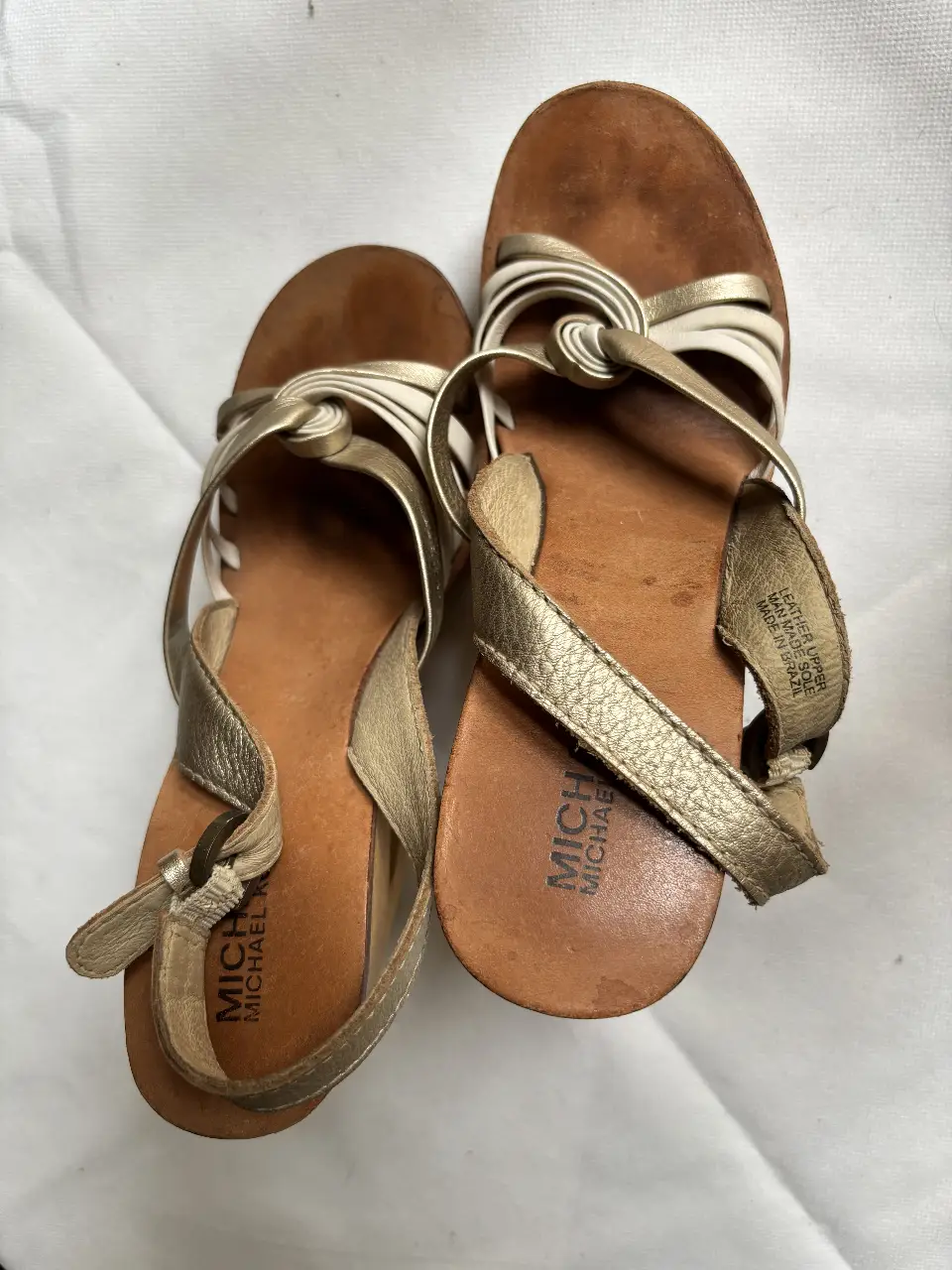 Sandalias Michael Kors doradas con detalles en blanco. Taco de madera y pulsera con hebilla para ajustar al tobillo. Ideales para un look elegante y cómodo.