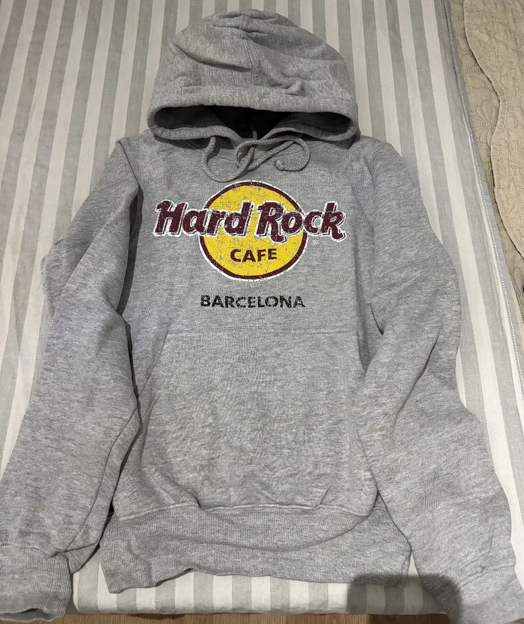 Buzo canguro gris de Hard Rock Cafe Barcelona, con estampado del logo en amarillo y borgoña. Talle XS, ideal para un look casual y urbano.