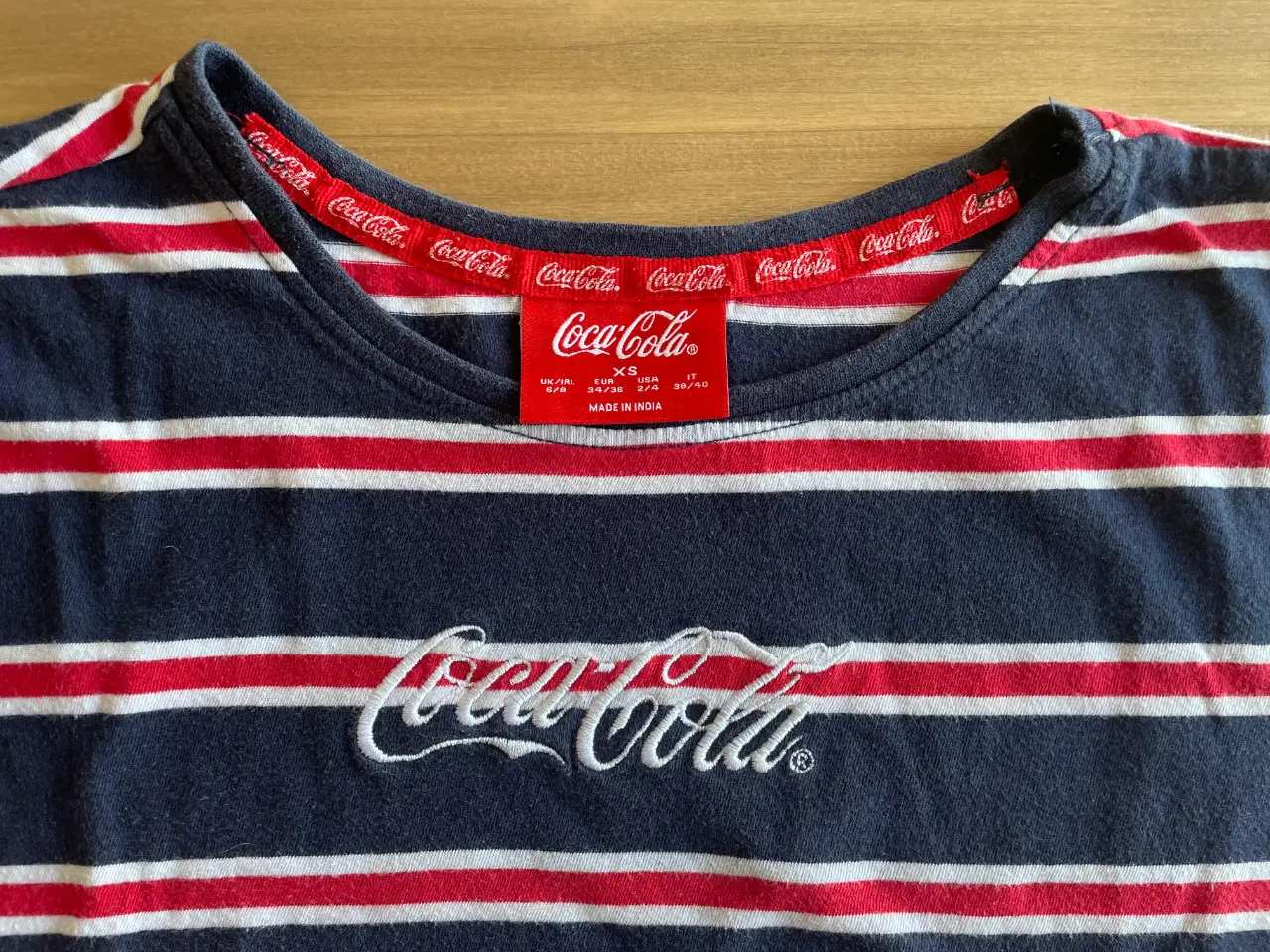 Remera de manga corta a rayas horizontales en azul marino, blanco y detalles en rojo. Con el logo de Coca-Cola bordado en el pecho y estampado en el cuello. Ideal para un look casual y cómodo. 46 cm Sisa, 39 cm largo.
