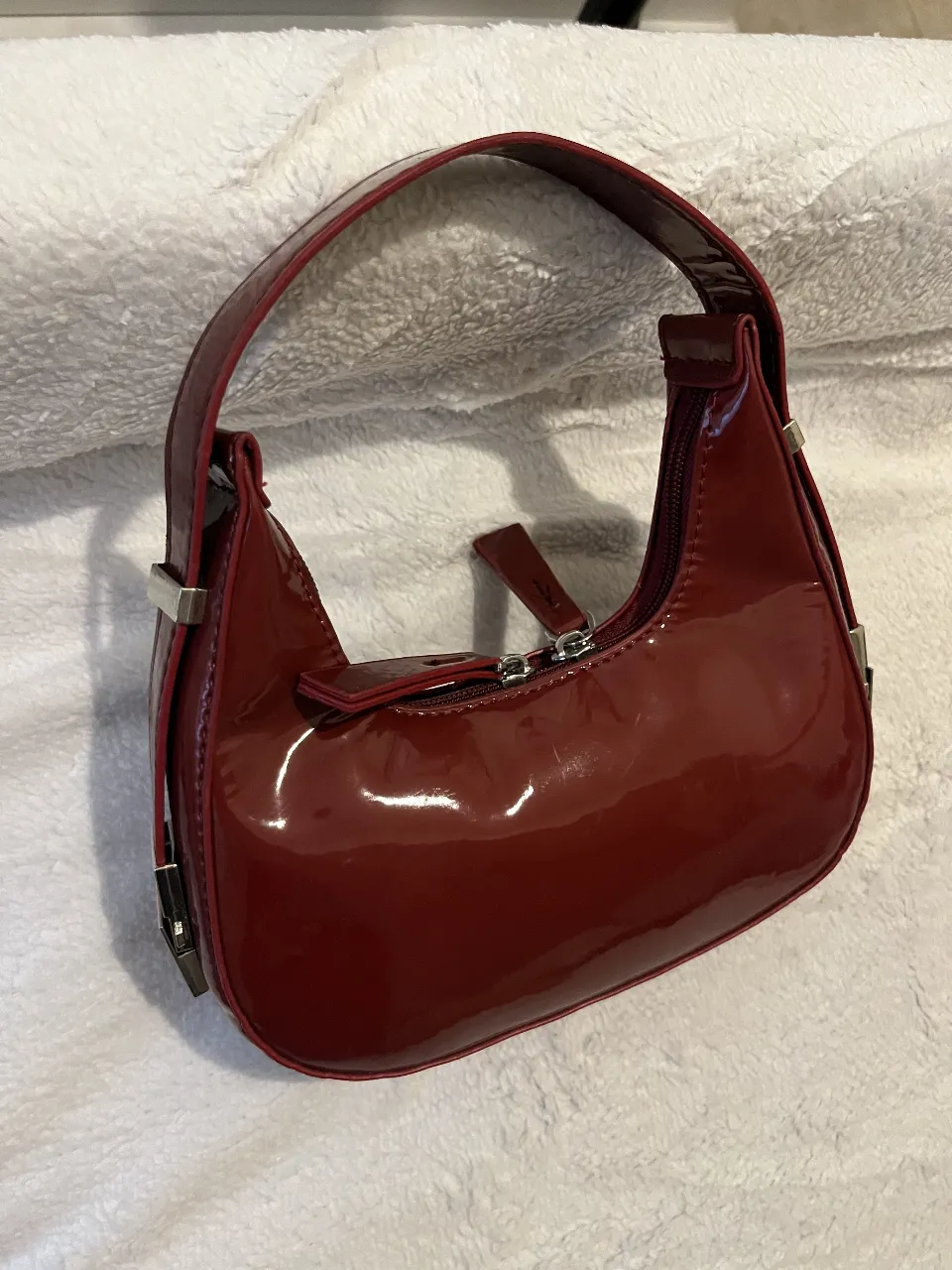 Cartera de charol color bordó, con correa de mano y cierre metálico. Ideal para darle un toque chic a cualquier outfit.