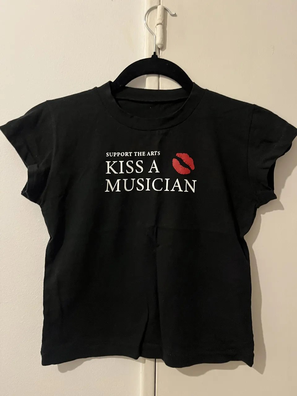 Remera negra corta con estampa "SUPPORT THE ARTS KISS A MUSICIAN" y un beso rojo. Ideal para un look casual y divertido.