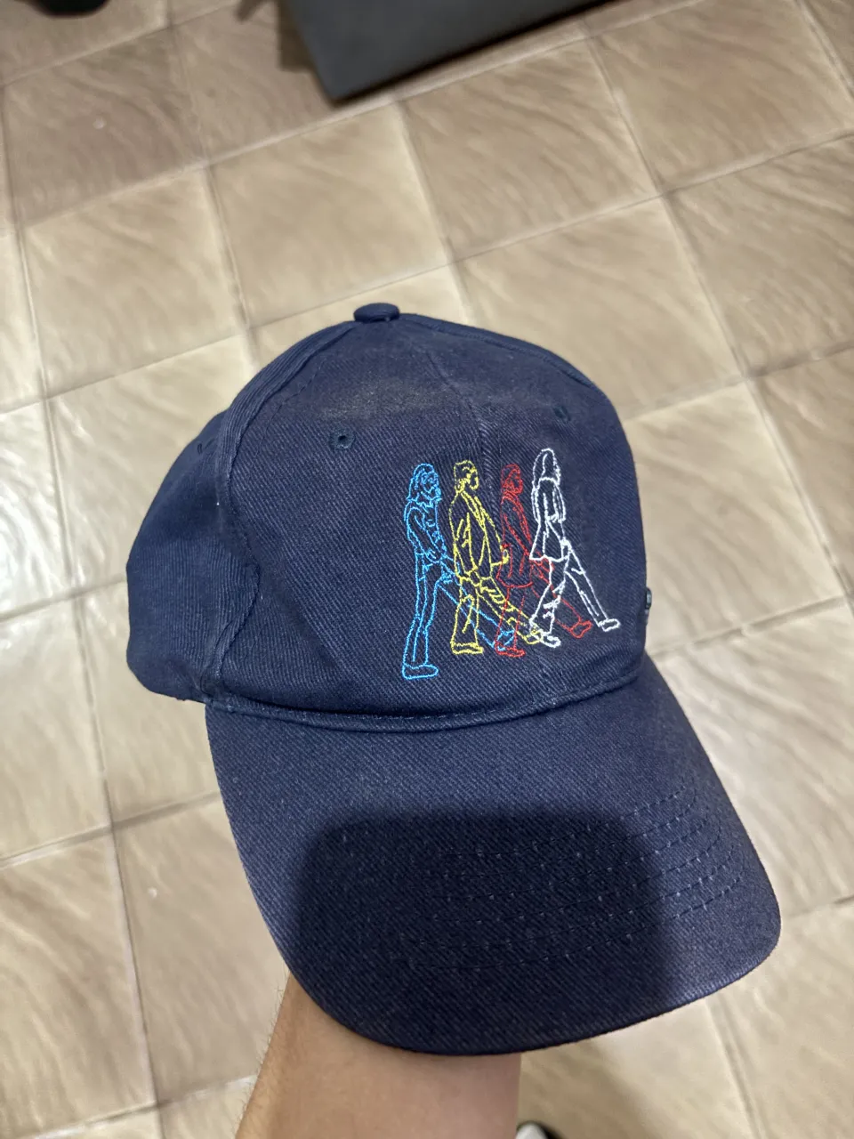 Gorra azul marino con bordado de los Beatles cruzando Abbey Road. Detalle de pin metálico lateral con el rostro de John Lennon. Ajuste regulable.