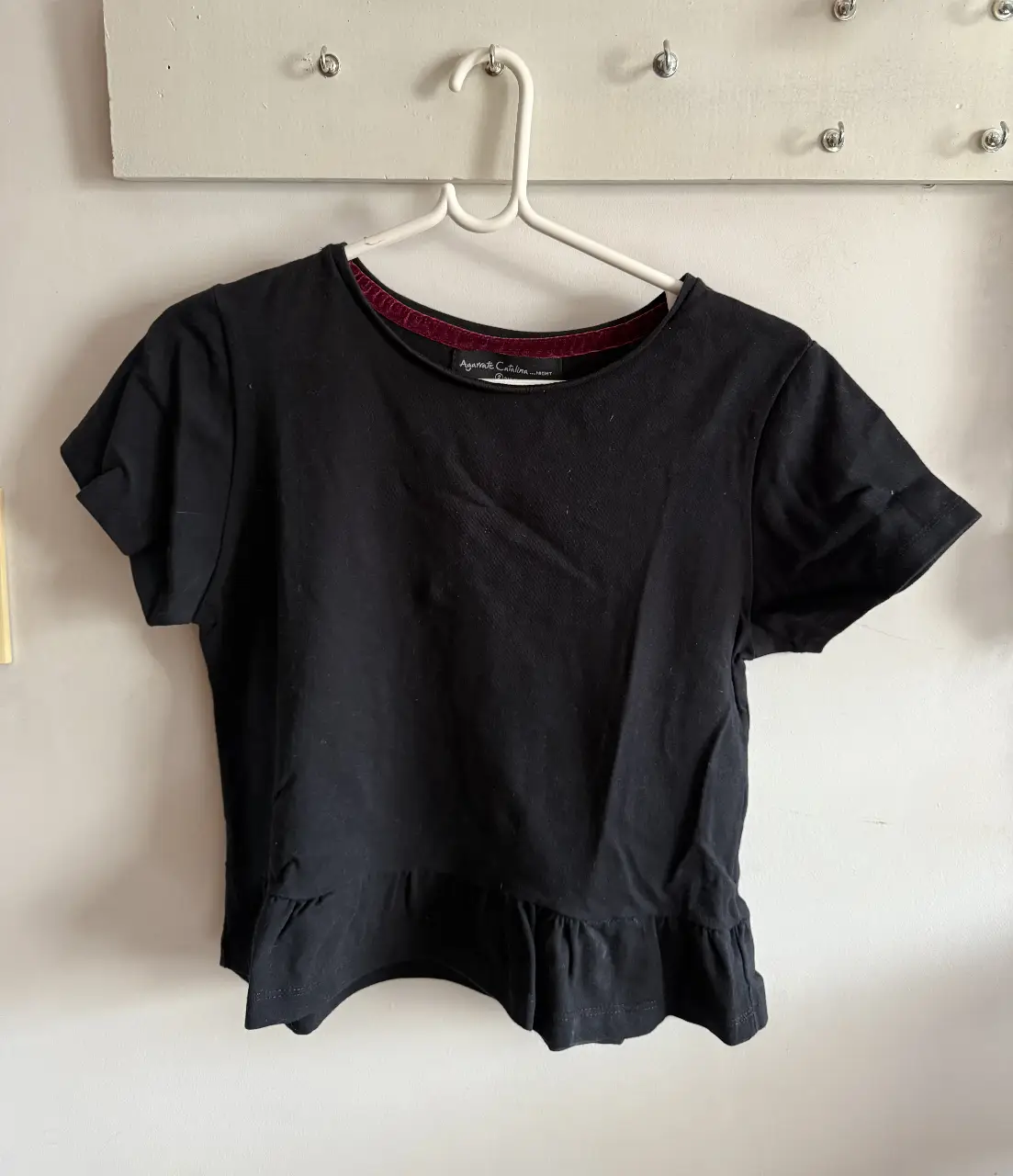 Remera negra de manga corta con detalle de volados en la parte inferior. Cuello redondo con borde en color borgoña. Ideal para un look casual y moderno.