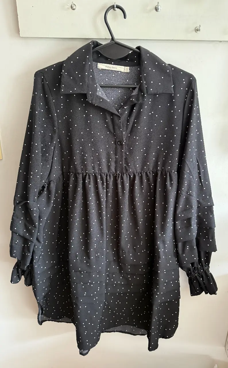 Blusa negra con lunares blancos, manga larga con volados y cuello camisero. Ideal para un look elegante y versátil.