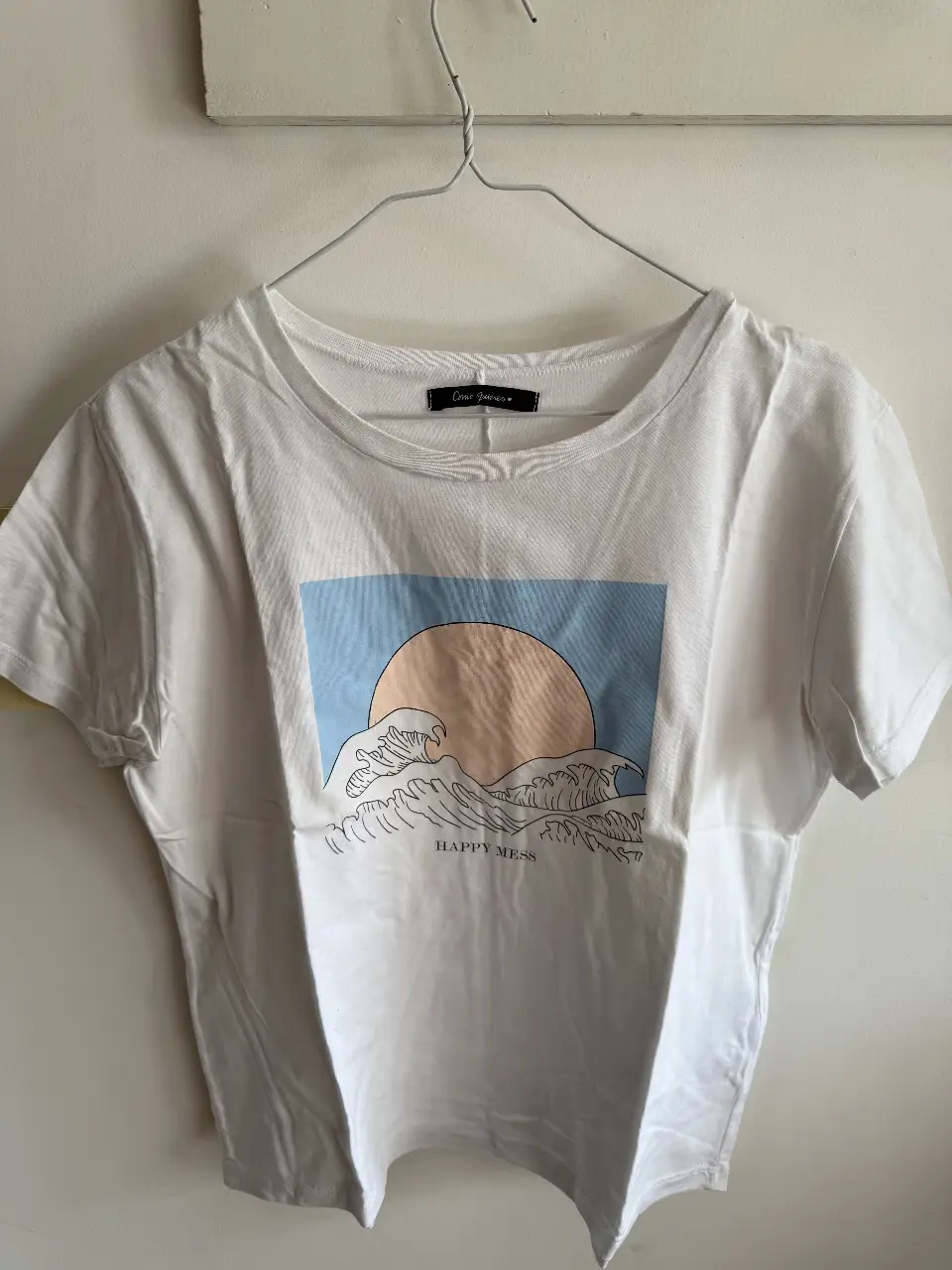Remera blanca de algodón con estampado de un sol y olas en tonos azules y beige, con la frase 'Happy Mess'. Ideal para un look casual y relajado.