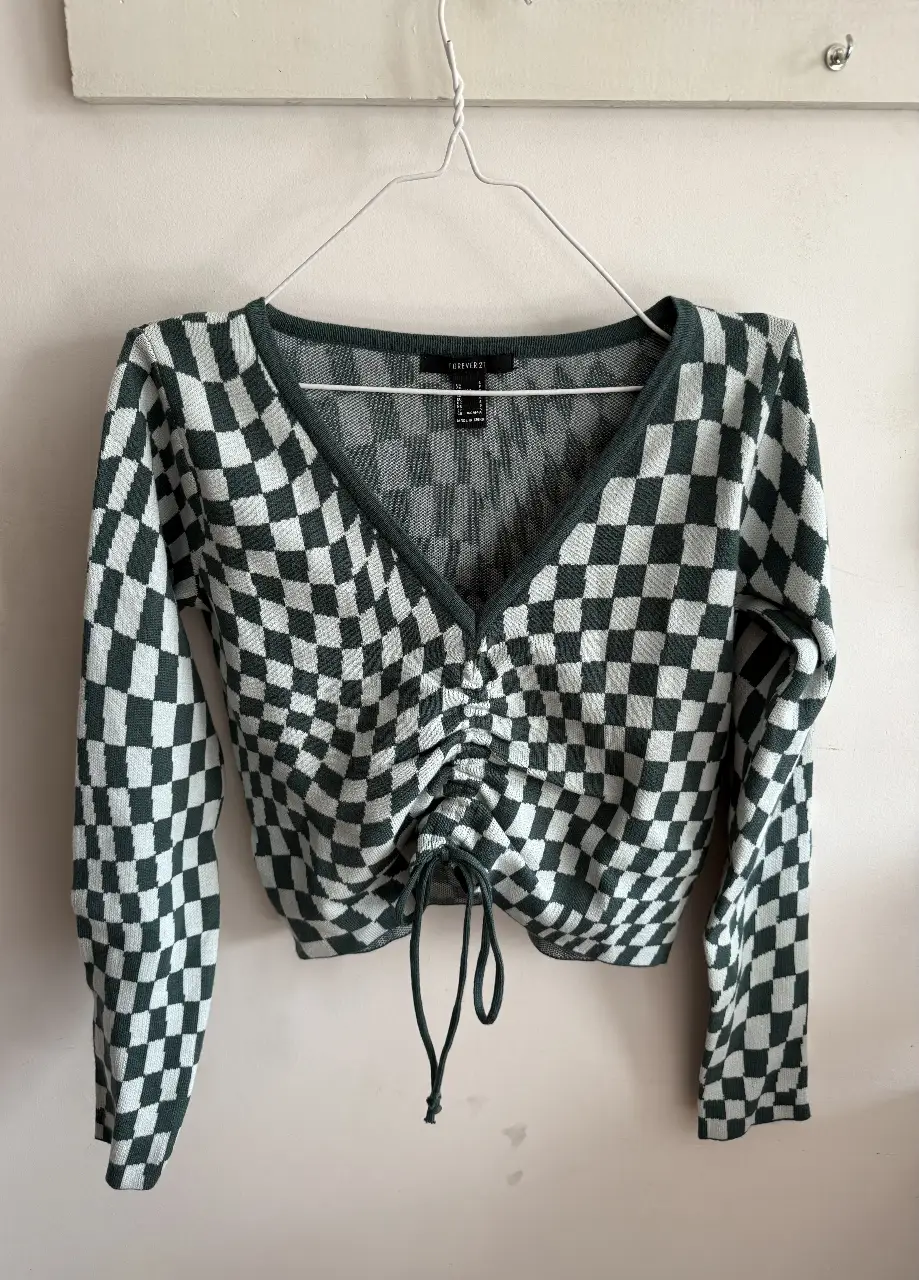Sweater de mujer con diseño a cuadros en verde y blanco. Tiene un escote en V y un detalle fruncido en el centro con cordones ajustables. Ideal para un look casual y moderno.
