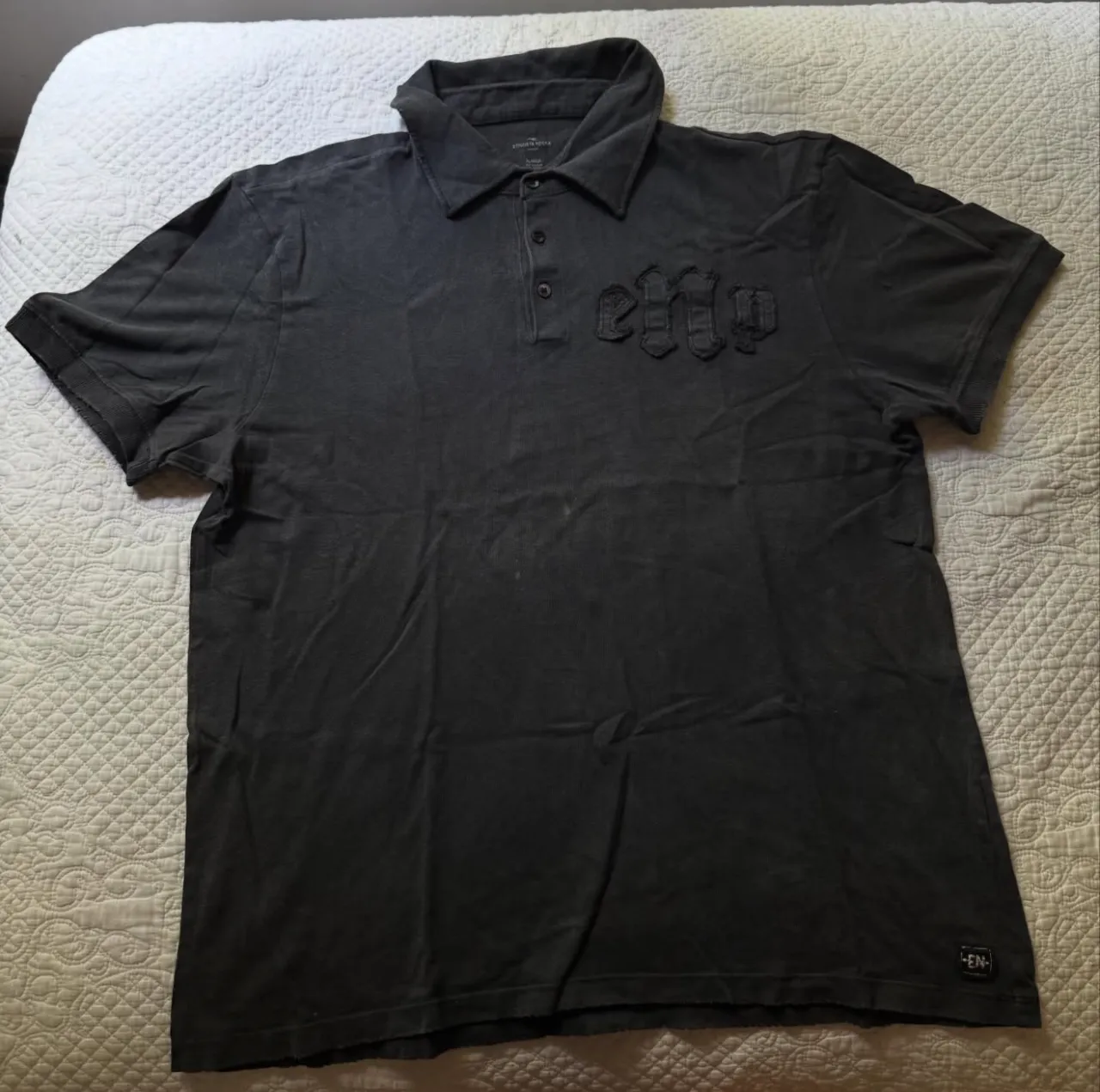 Remera tipo polo gris oscuro, con cuello y mangas cortas. Tiene un diseño en relieve en la parte delantera y un pequeño logo en la manga.