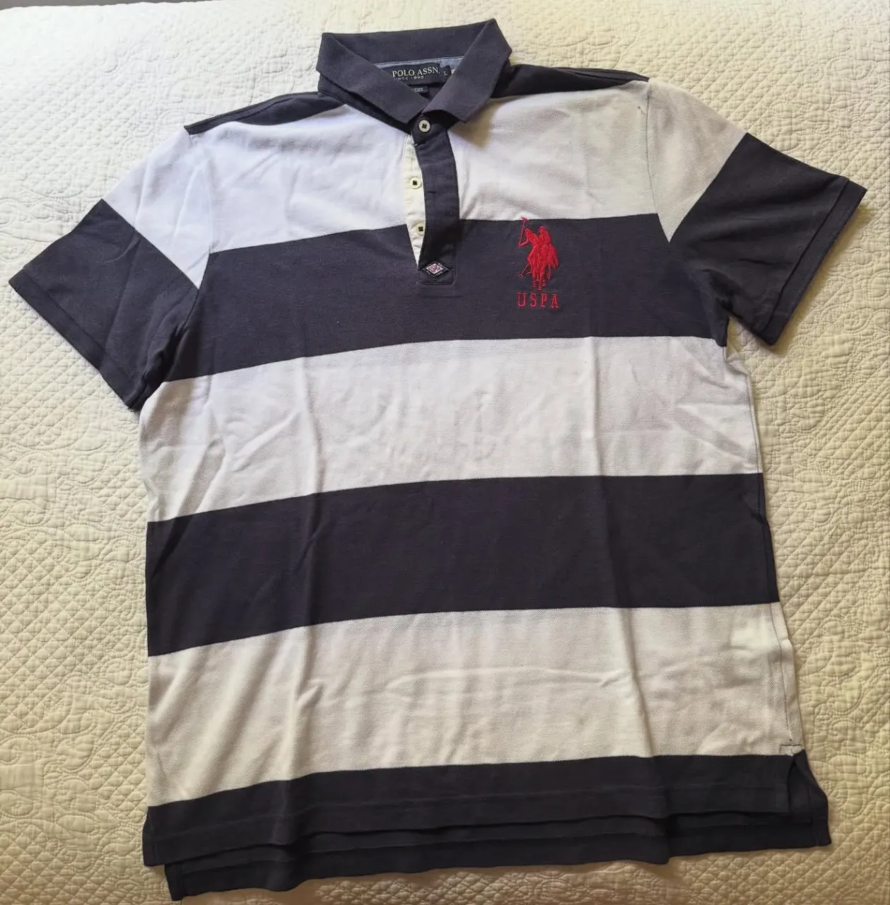 Remera polo a rayas grises y blancas, con logo bordado de "USPA" en rojo. Ideal para un look casual y cómodo.