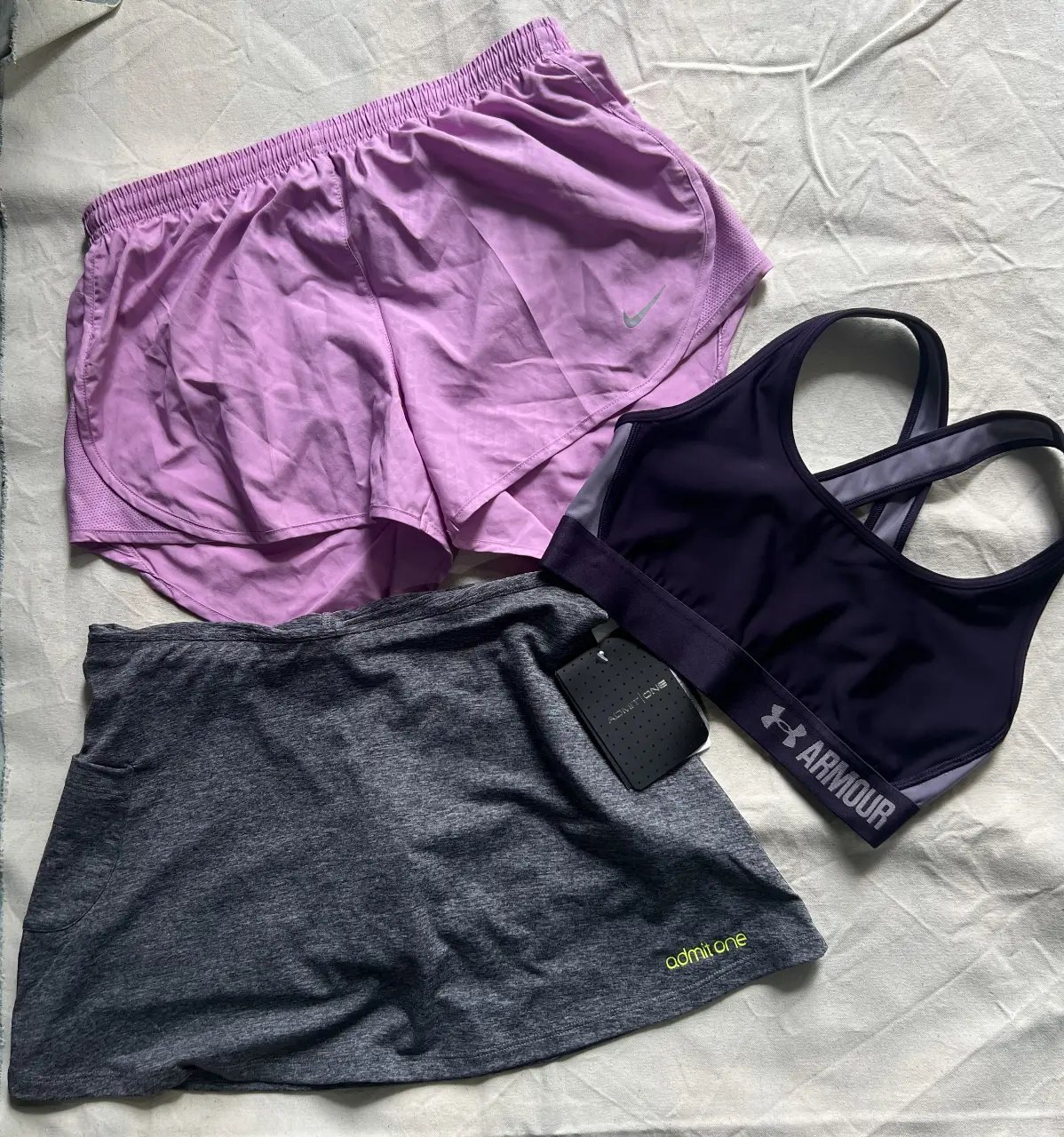 Conjunto deportivo de mujer, incluye shorts Nike color violeta con logo y top deportivo Under Armour color violeta oscuro. Además, una pollera short gris Admit One con etiqueta. TODO TALLE SMALL