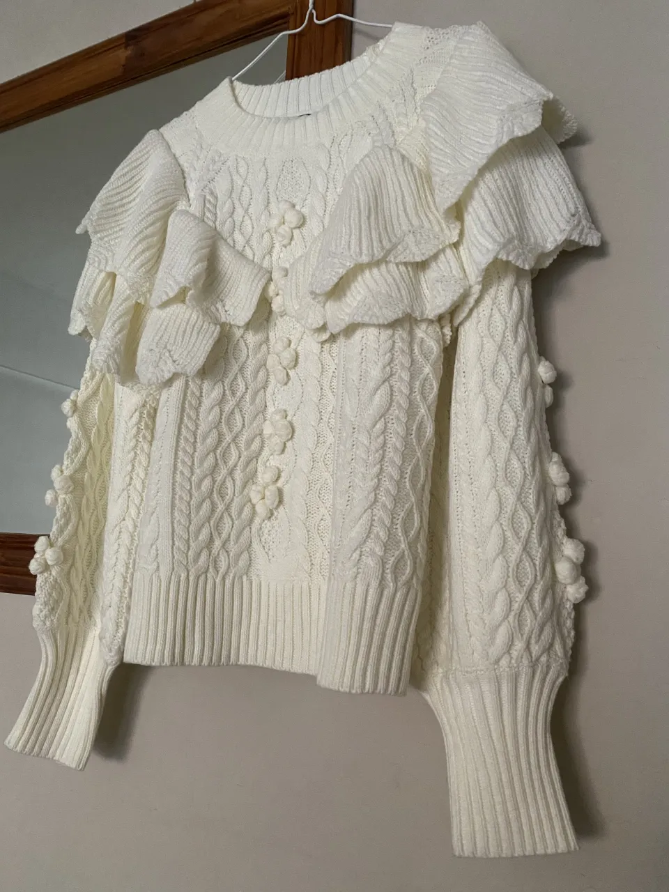 Sweater blanco de tejido con trenzas y detalles de pompones. Posee volados en la parte superior del busto y mangas largas con puños ajustados.