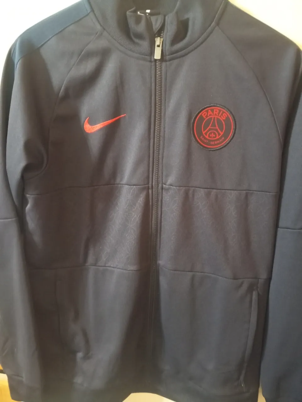 Buzo deportivo con cierre, de color azul marino con detalles en rojo. Cuenta con el logo de Nike y el escudo del Paris Saint-Germain. Ideal para usar en actividades deportivas o para un look casual.