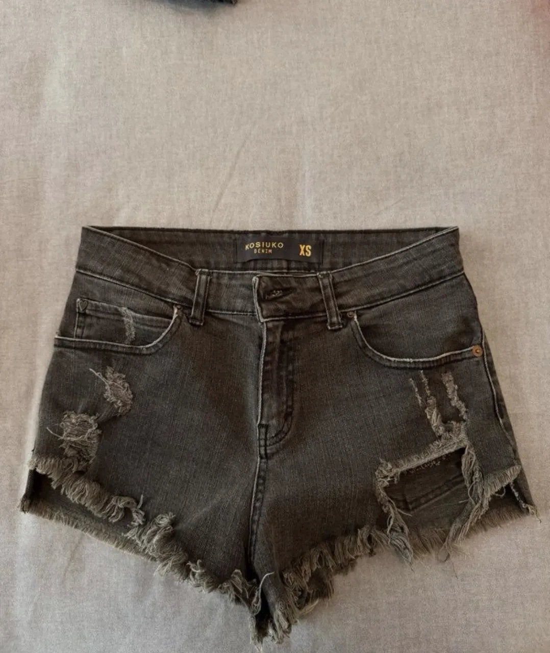 Shorts de jean grises con roturas y detalles deshilachados en los bordes inferiores y en los bolsillos delanteros. Detalle de estrella dorada en el bolsillo trasero derecho.