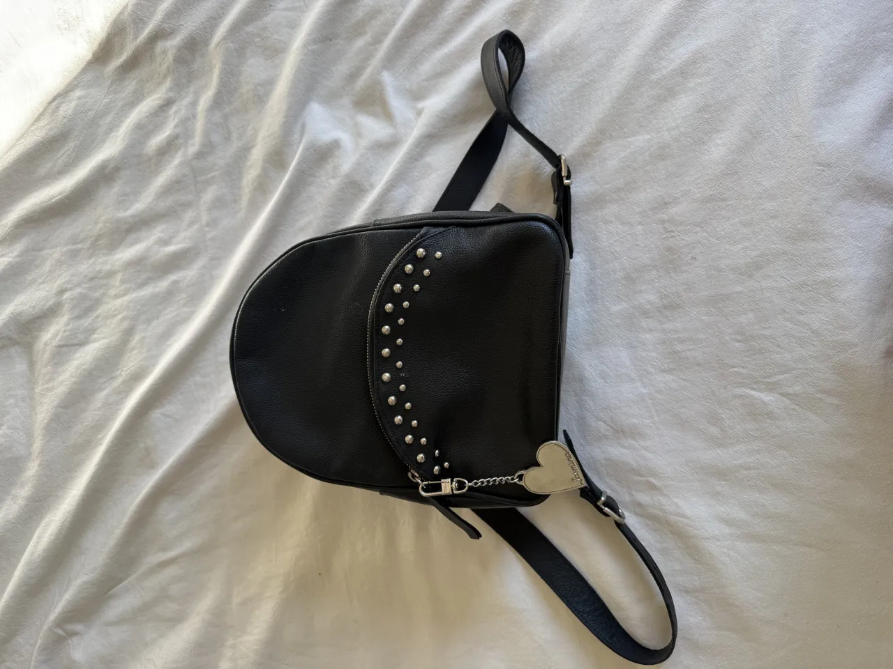 Mochila negra de cuero sintético con tachas plateadas y un charm de corazón colgante. Ideal para darle un toque rockero y canchero a tu look.