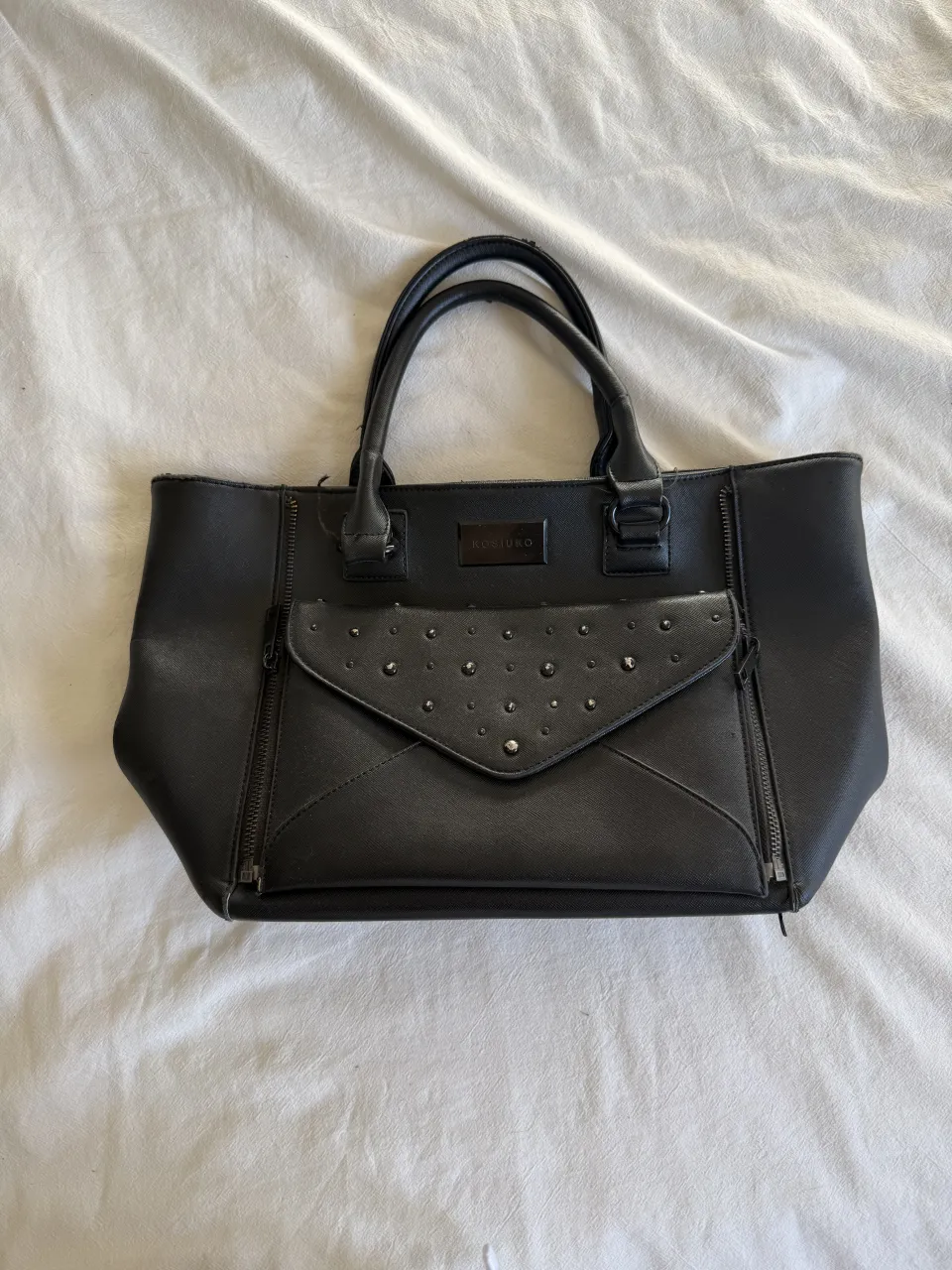 Bolso negro de cuero sintético, con tachas y bolsillo frontal. Ideal para el día a día o para salir.