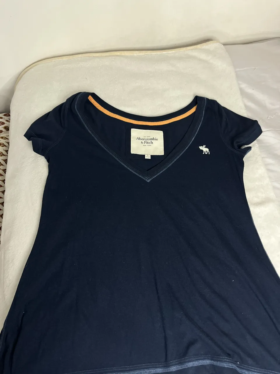 Remera azul marino de Abercrombie & Fitch con escote en V y detalle de ribetes en naranja. Tiene un pequeño bordado de alce en el frente. Ideal para un look casual y cómodo.