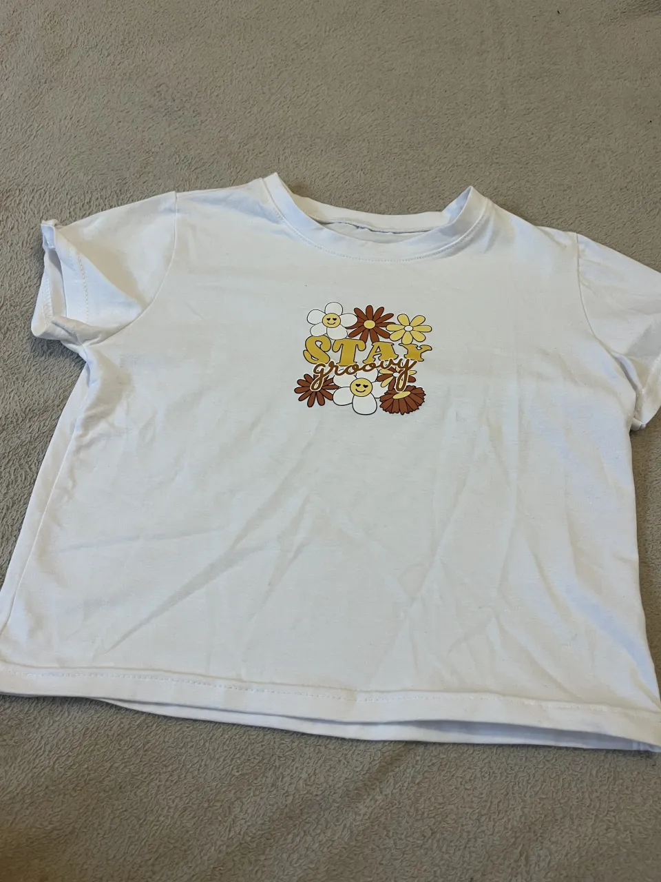 Remera blanca de manga corta con estampa "Stay groovy" rodeada de flores y caritas sonrientes.