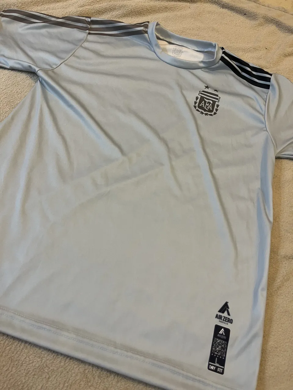 Remera de manga corta celeste con detalles grises en los hombros y el cuello. Presenta un escudo de la AFA estampado en el frente 