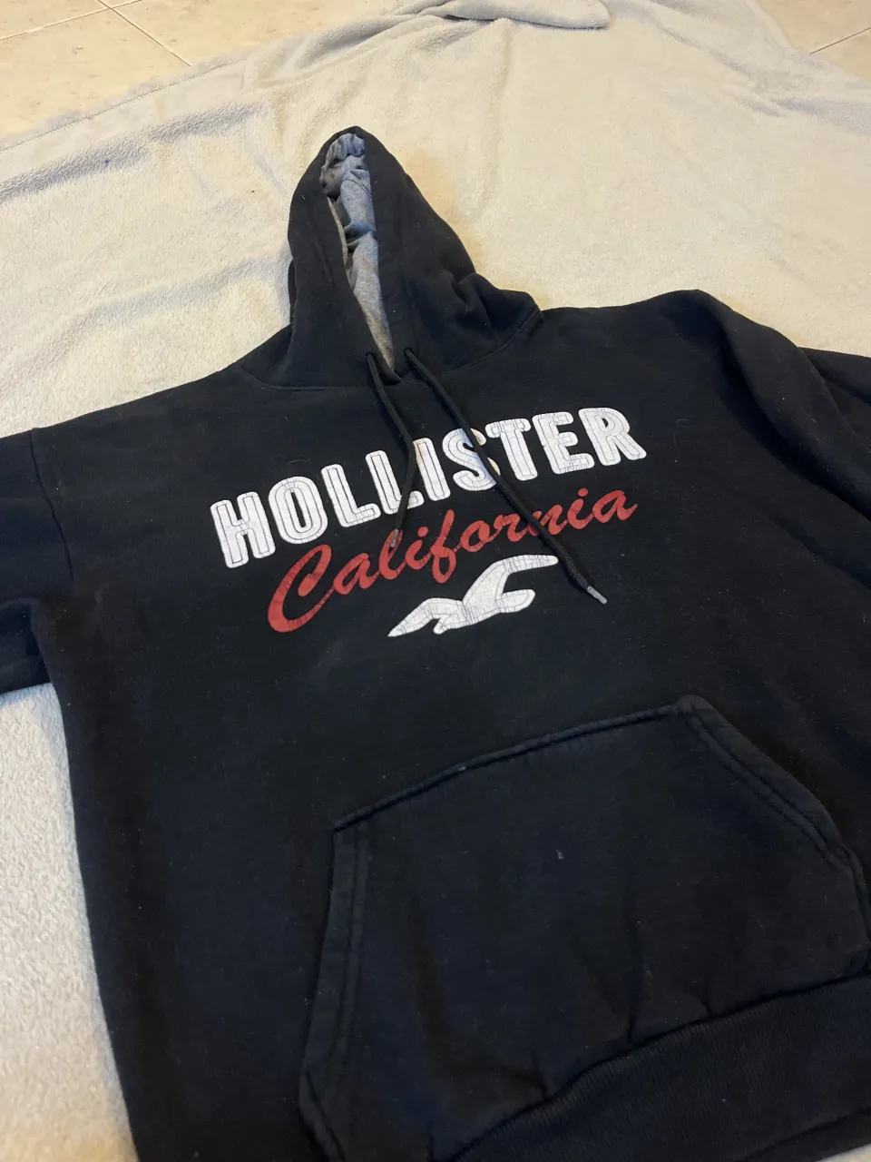 Buzo negro con capucha y cordones. Estampado frontal con el logo "HOLLISTER California" en blanco y rojo. Posee bolsillo canguro. Aprox un talle M