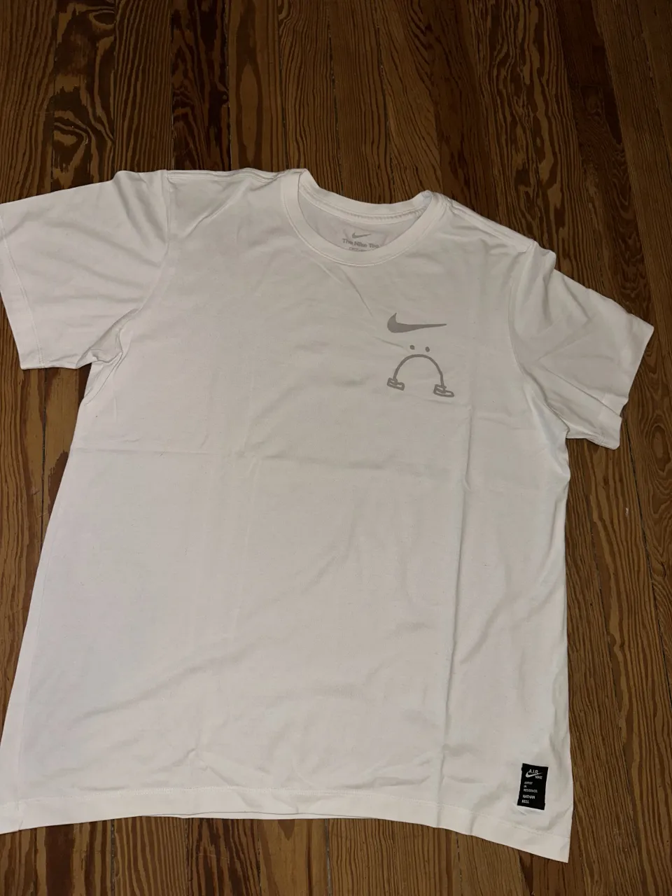 Remera blanca de manga corta con estampa gráfica en el frente y en la espalda. La estampa en la espalda dice "I CAN'T FEEL MY LEGS" con un dibujo. Tiene un logo de Nike y una etiqueta "A.I.R. NIKE ARTIST IN RESIDENCE: NATHAN BELL" en la parte inferior del frente.