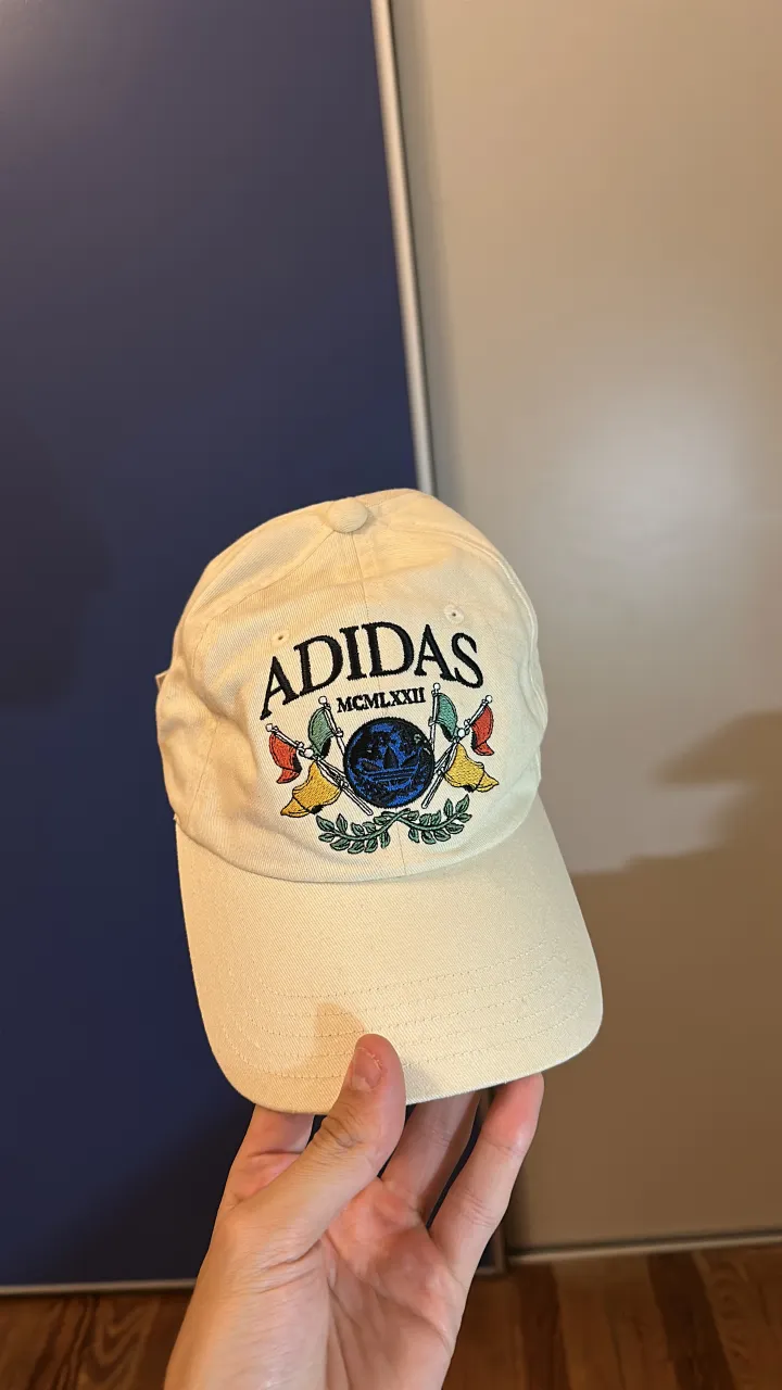 Gorra de algodón color crema de Adidas, con un diseño bordado en el frente que incluye el logo de la marca, el año "MCMLXXII" y un globo terráqueo con banderas. Es un accesorio urbano y canchero, ideal para complementar looks casuales.