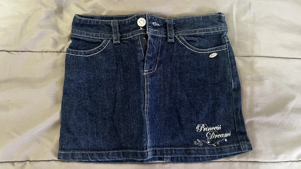 Pollera de jean azul oscuro con bordado "Princess Dreams" y detalles de costura blanca. Cierre y botón.