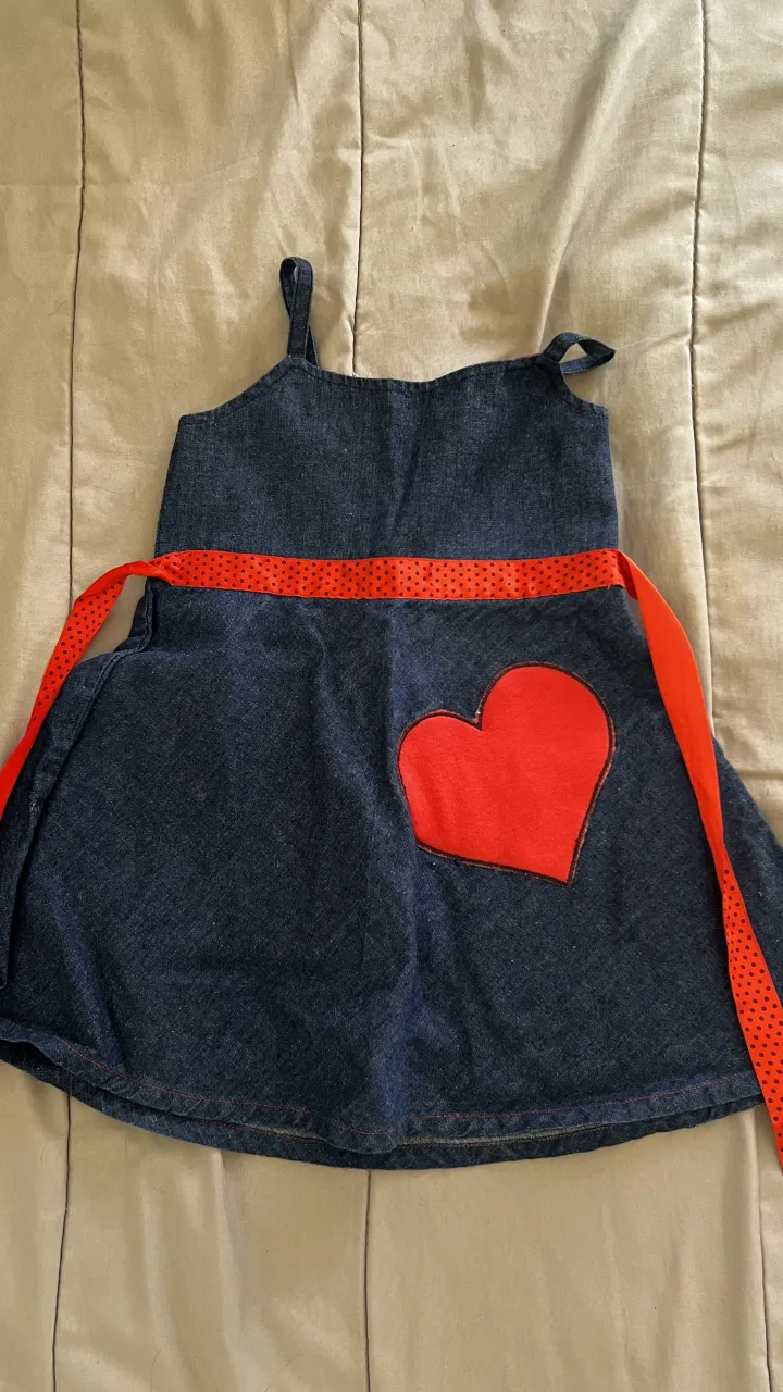 Vestido de jean azul con cinturón rojo y estampa de corazón rojo en el frente. Tiene tiradores finos y cierre en la espalda.