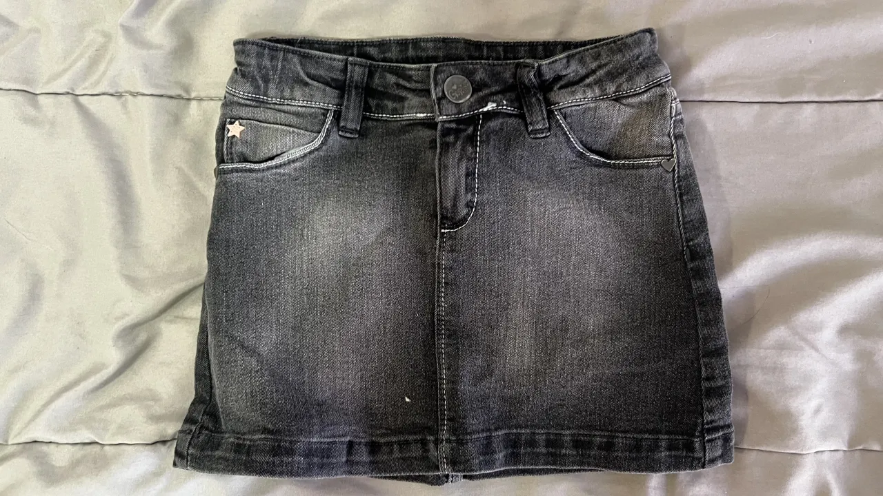 Pollera de jean gris oscuro, con detalles de costuras blancas. Tiene un bolsillo delantero con una estrella bordada y bolsillos traseros, uno de ellos con un corazón aplicado.