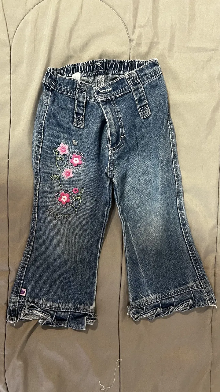 Jean de bebé con elástico en la cintura y detalle de flores bordadas en la pierna izquierda. La parte inferior de las piernas tiene un volante.