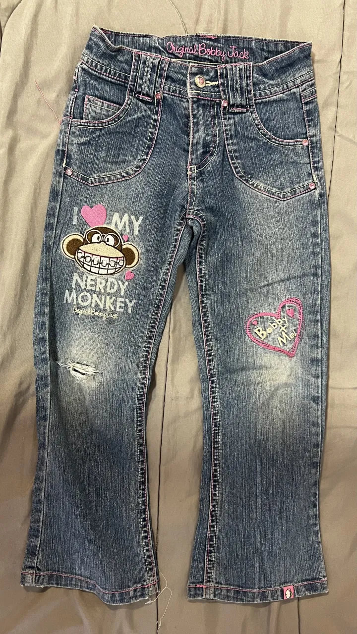 Jean de nena con bordados de mono y detalles de corazón. Presenta rotura en la rodilla izquierda y detalles de costura rosa en los bolsillos y dobladillos.