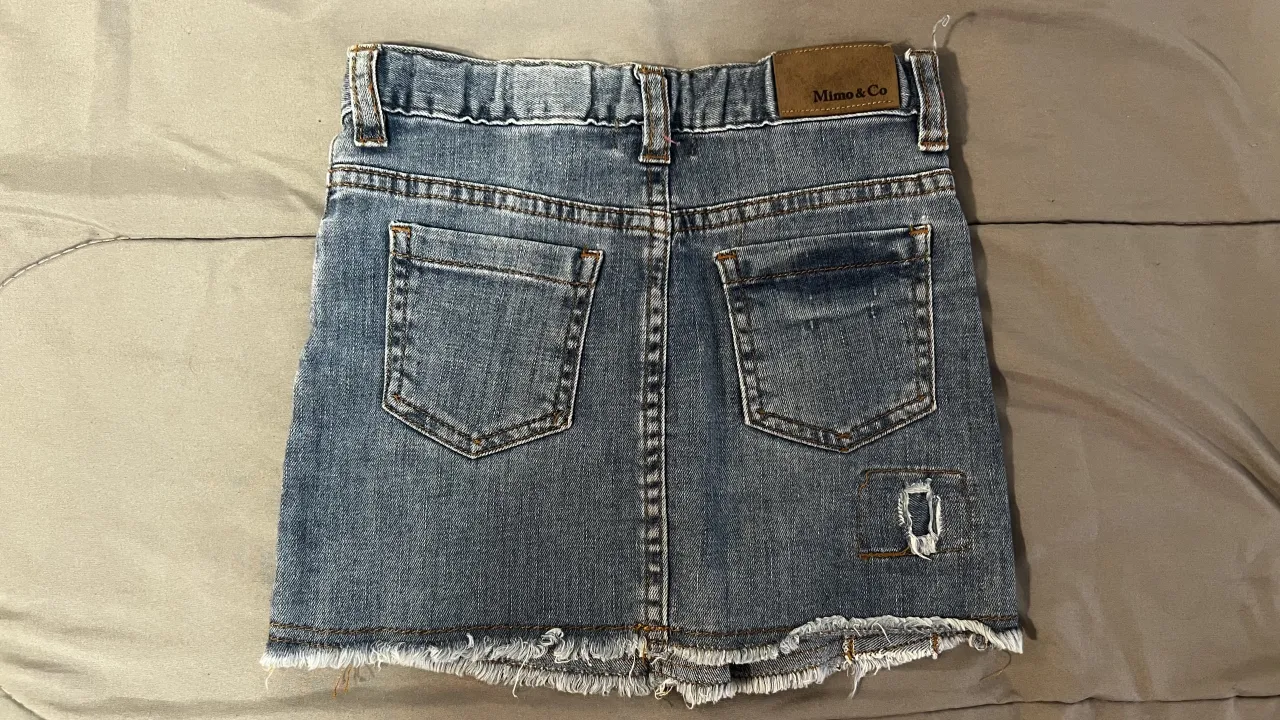 Pollera de jean con roturas y detalles deshilachados en el borde inferior. Presenta parches de tela con roturas en la parte delantera y trasera.