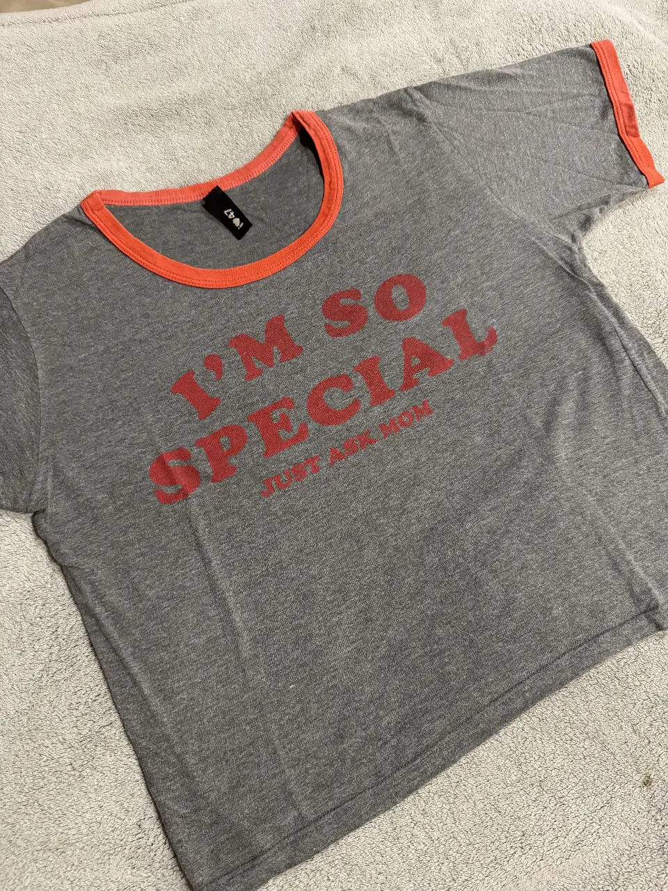 Remera gris con cuello y mangas en color naranja. Estampado "I'M SO SPECIAL JUST ASK MOM" en rojo. Ideal para un look casual y divertido.
