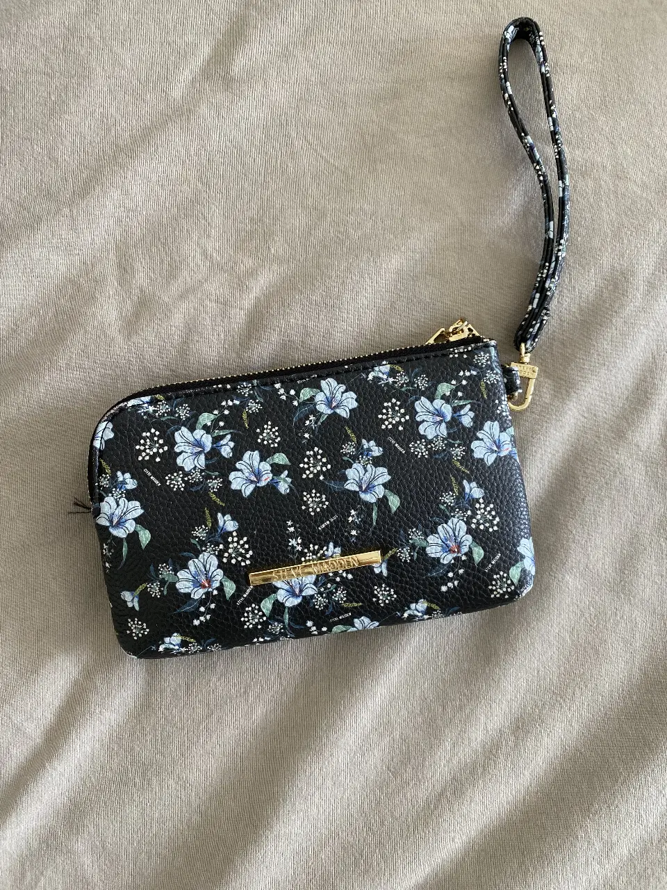 Monedero/Billetera Steve Madden de mano con estampado floral en tonos azules y blancos sobre fondo negro. Cuenta con cierre dorado y correa para la muñeca. Ideal para llevar lo esencial con estilo.