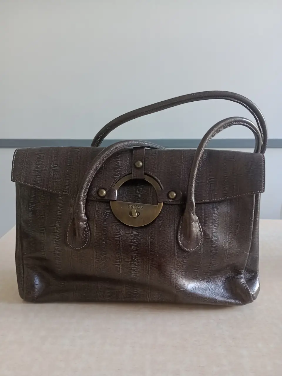 Cartera de mano PRÜNE de cuero marrón con detalles metálicos dorados. Diseño elegante y sofisticado, ideal para complementar cualquier look.