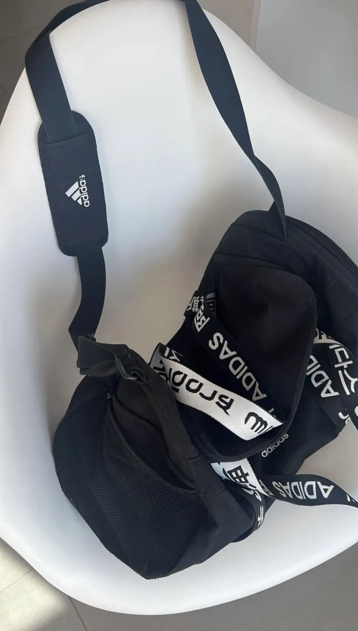 Bolso deportivo negro con correa ajustable y logo de Adidas repetido en la correa principal. Posee múltiples compartimentos y cierres.