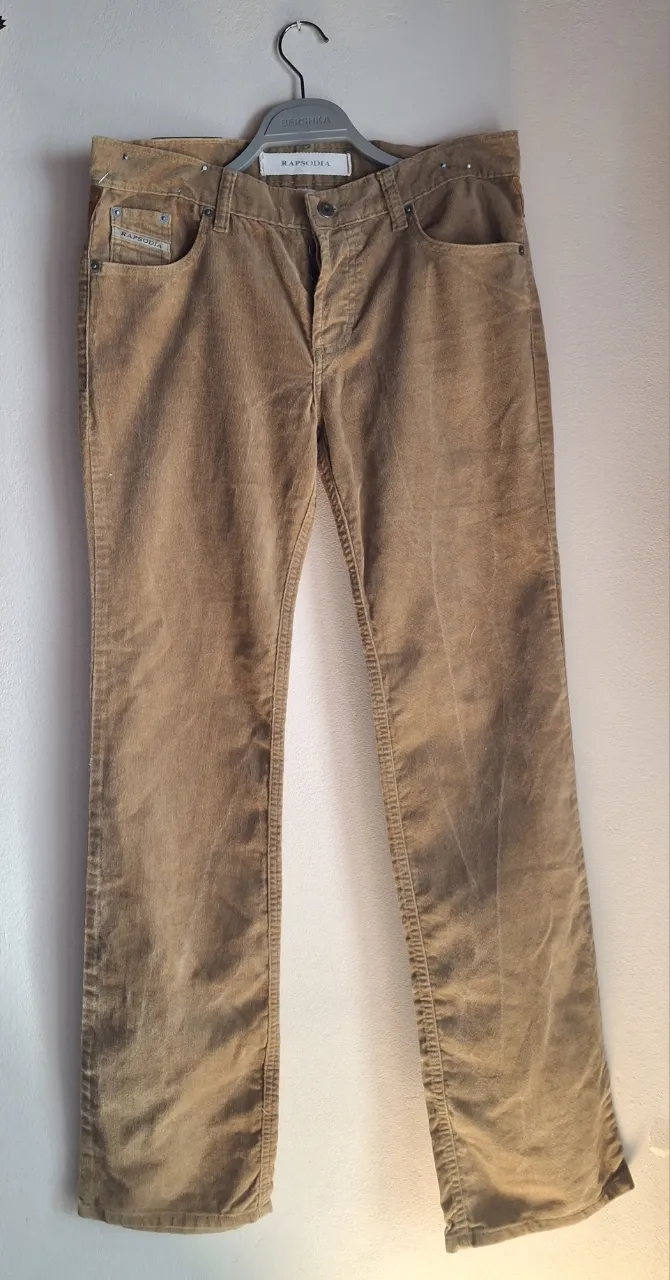 Pantalón de pana color marrón claro, tiro bajo, con detalles de tachas en la cintura y un cinturón de cuero con apliques metálicos. Posee bolsillos delanteros y traseros. Ideal para un look casual y canchero.