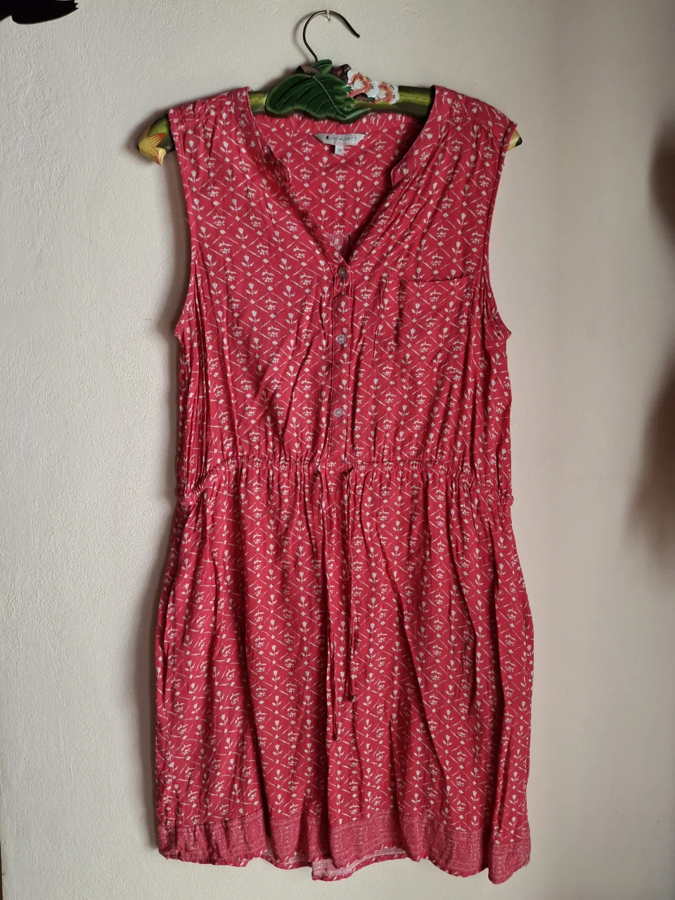 Vestido corto de verano, sin mangas, con un estampado floral pequeño en color rosa vibrante. Posee un cuello en V con botones y un cordón ajustable en la cintura para un calce cómodo y favorecedor. Ideal para días soleados y salidas informales.