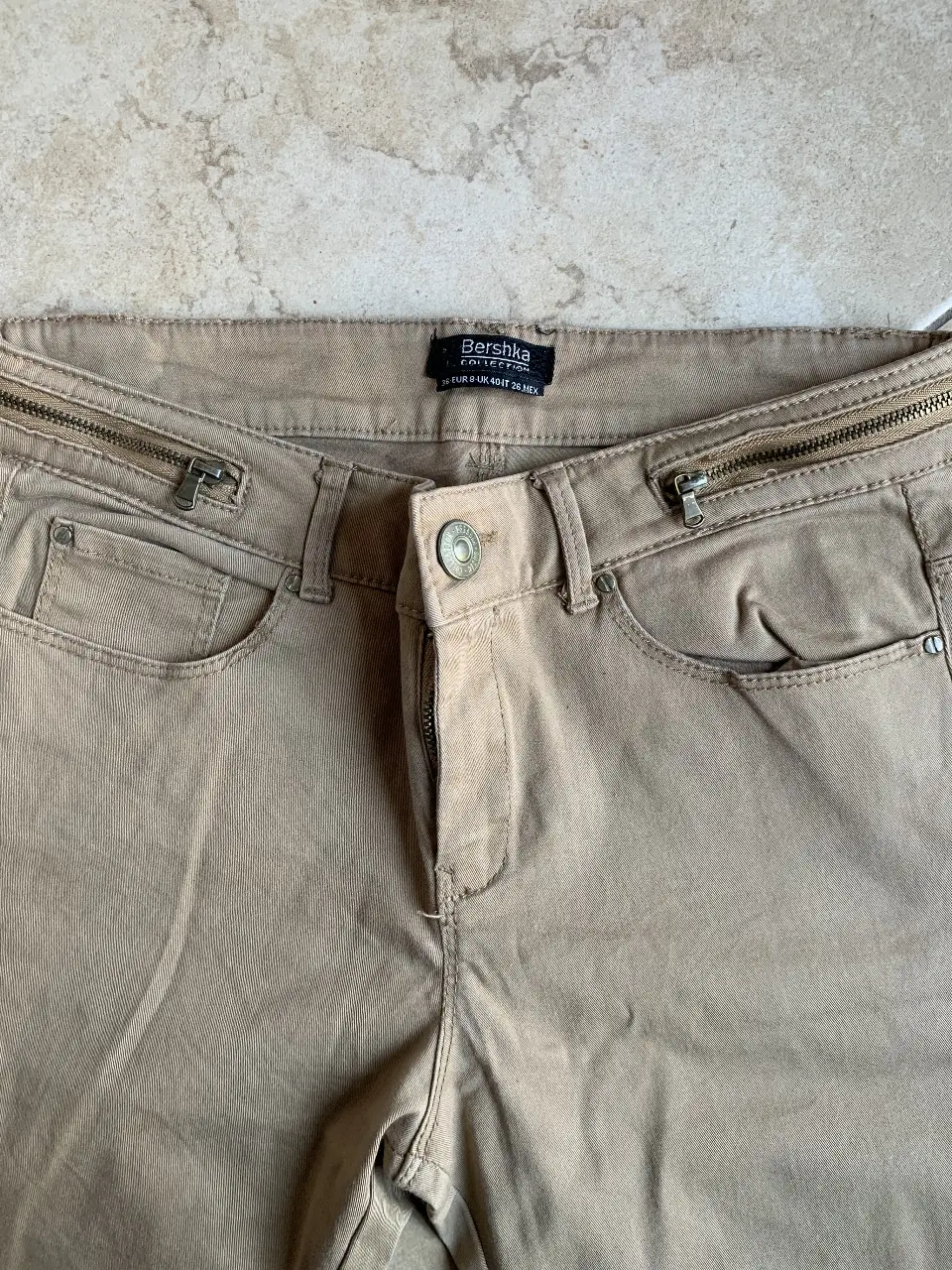 Pantalón de vestir color khaki de la marca Bershka, con cierre y detalles de cremalleras en la cintura. Ideal para un look casual y moderno.