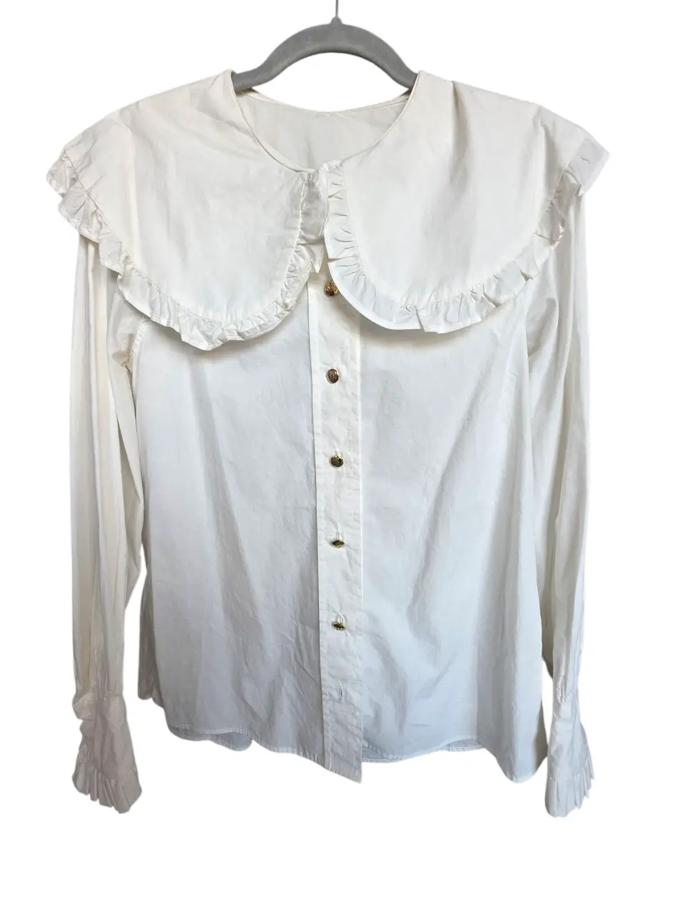 Camisa blanca de algodón. Cuello amplio con voladitos y botones dorados. Mangas largas, puños con volados y botón. 