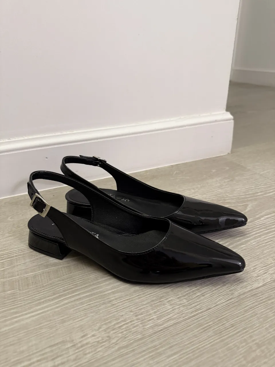 Zapatos negros de charol con punta fina y tiras al talón. Tienen un taco bajo y una hebilla para ajustar. Ideales para un look elegante y moderno.