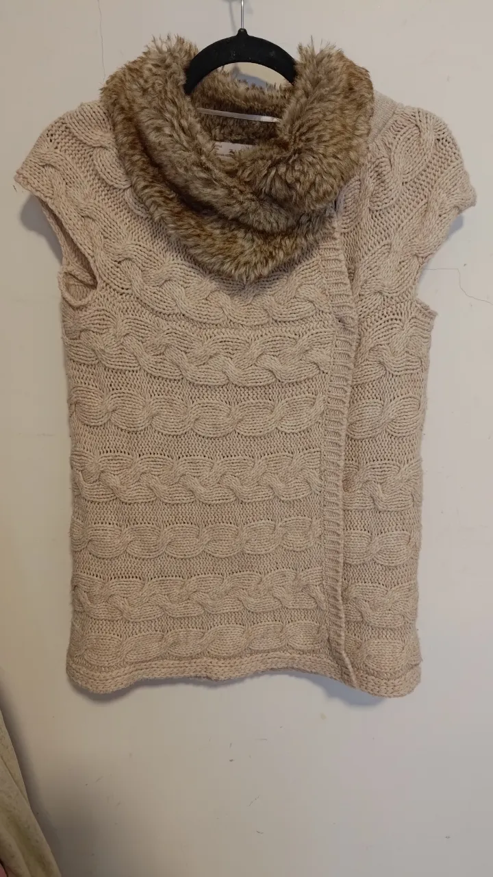 Chaleco tejido color crudo con cuello de piel sintética. Posee botones metálicos y un diseño de trenzas muy canchero. Ideal para un look abrigado y con estilo. talle m