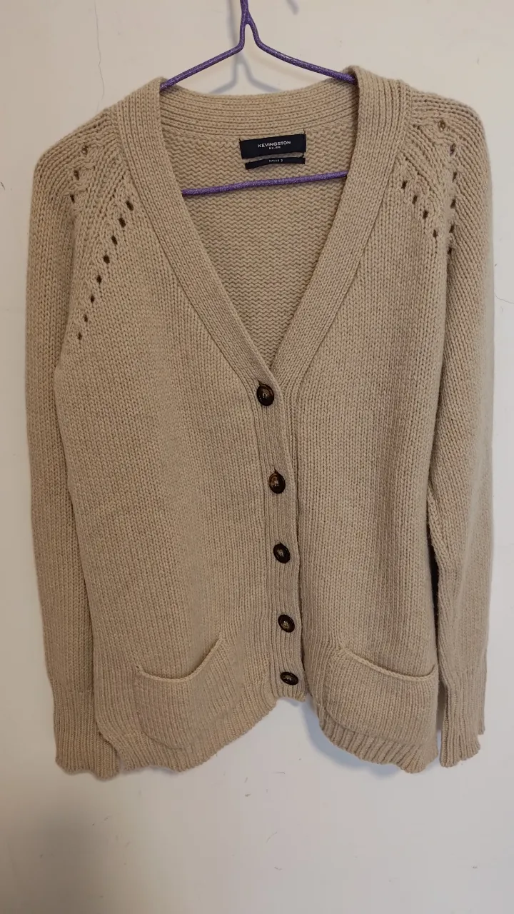 Cardigan de tejido de punto color tiza, con botones marrones y bolsillos delanteros. Posee detalles calados en los hombros que le dan un toque delicado y moderno. Ideal para un look casual y cómodo.