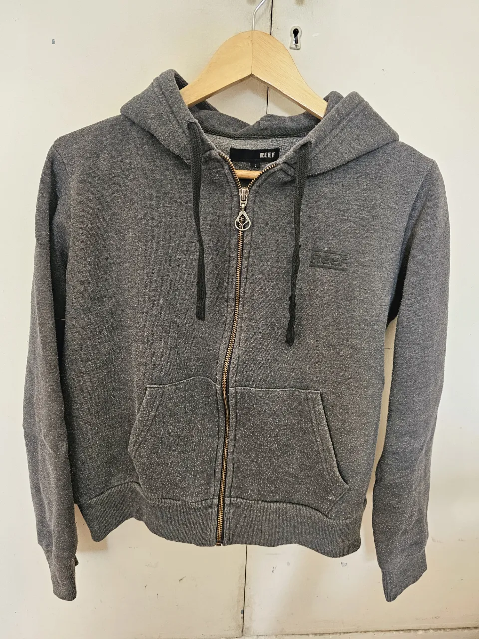 Buzo con cierre y capucha de color gris, con logo de la marca REEF en el pecho. Ideal para un look casual y cómodo.