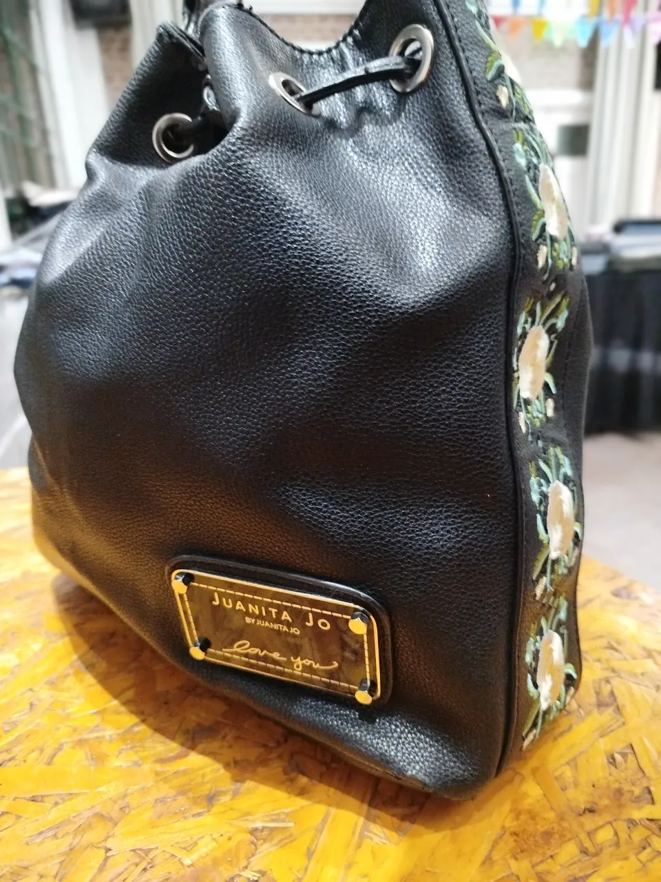 Cartera tipo balde de cuero negro con detalles florales bordados en un lateral. Cuenta con un cierre de ajuste con cordón y una placa dorada con el nombre de la marca. Ideal para un look casual y moderno.