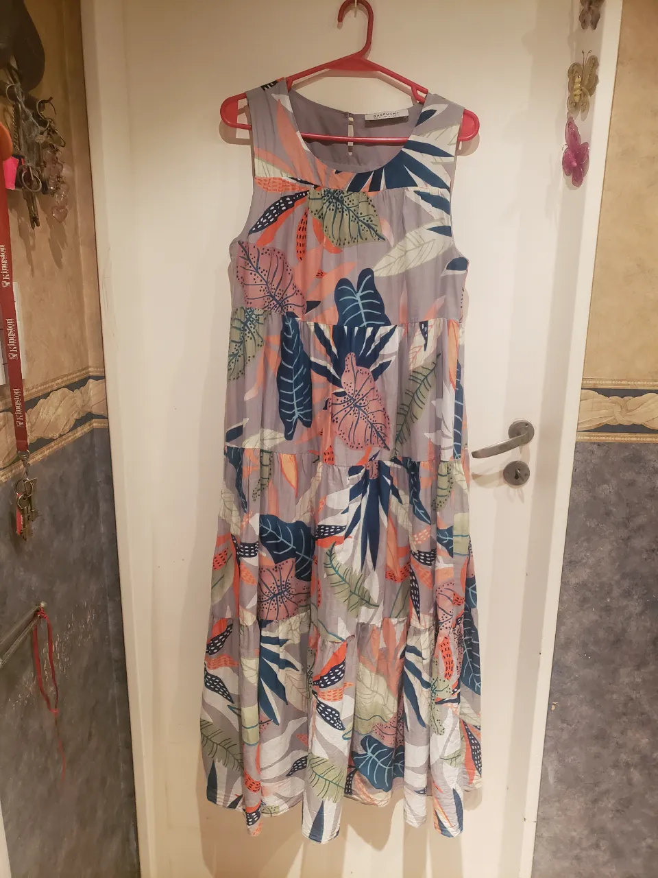 Vestido largo estampado con hojas y flores en tonos grises, azules, rosas y naranjas. Diseño con volados y escote redondo. Ideal para un look fresco y moderno.