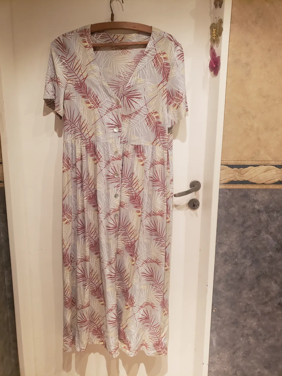 Vestido largo estampado de hojas en tonos rojos, amarillos y blancos sobre fondo gris. Posee cuello en V y botones en la parte superior. Ideal para un look casual y fresco.