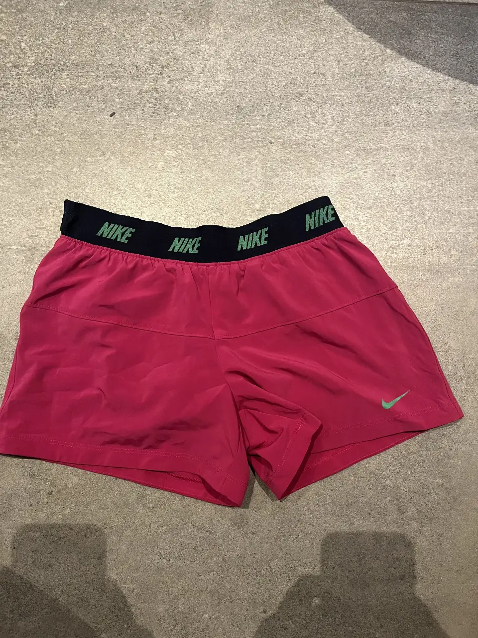 Short deportivo Nike Dri-Fit rosa para mujer, con cintura elastizada negra con logo de la marca en verde. Ideal para entrenamientos y actividades físicas.