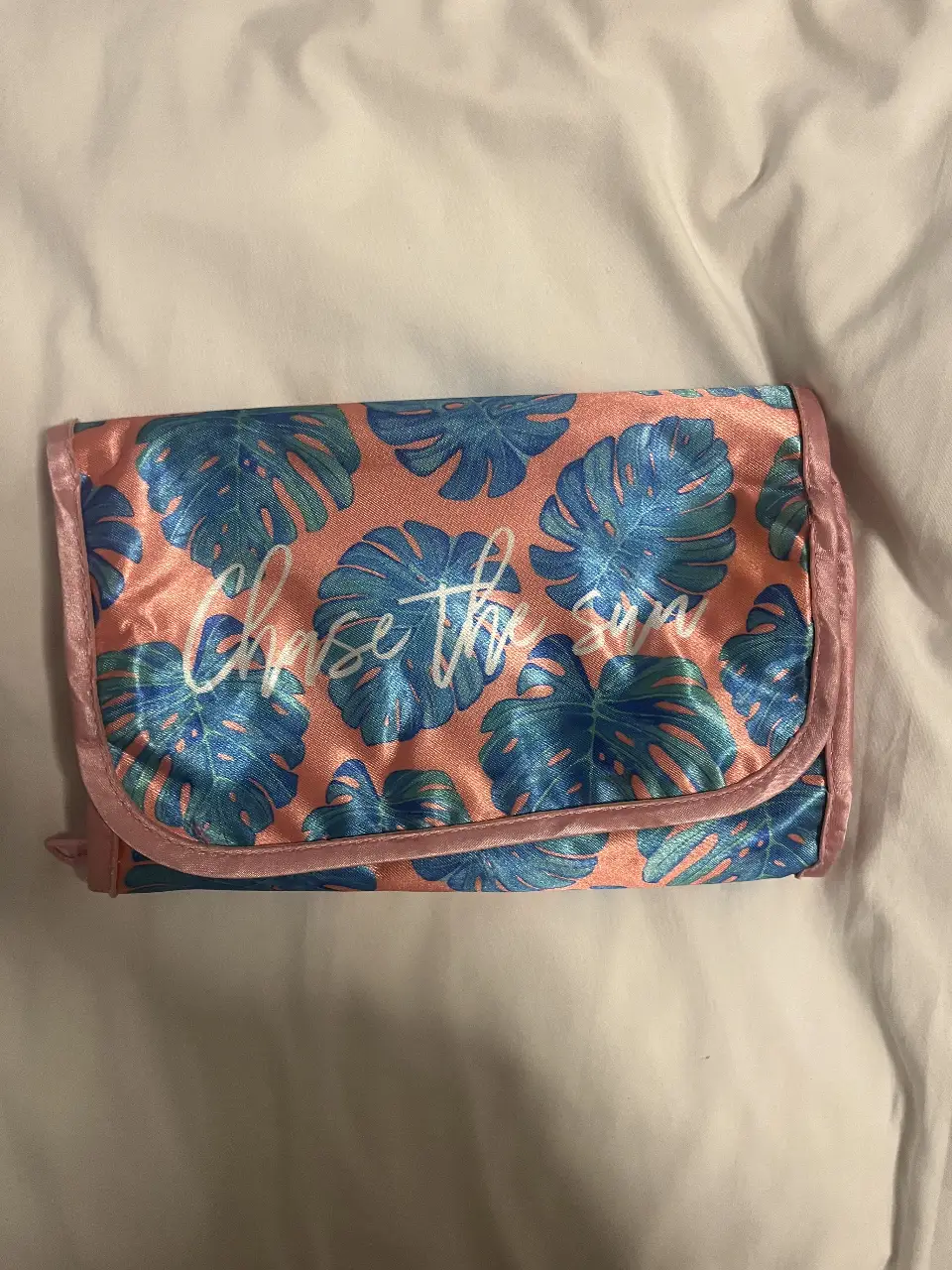 Neceser de viaje con estampado de hojas tropicales en tonos rosa y azul. Ideal para guardar cosméticos y artículos de aseo personal. Cuenta con varios compartimentos y cierres para organizar tus pertenencias.