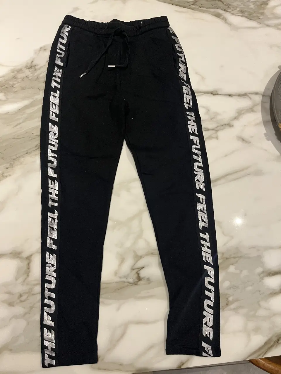 Pantalón deportivo negro con detalles en blanco. Tiene la frase "THE FUTURE FEEL THE FUTURE" en letras blancas a lo largo de los laterales. Ideal para un look casual y cómodo.
