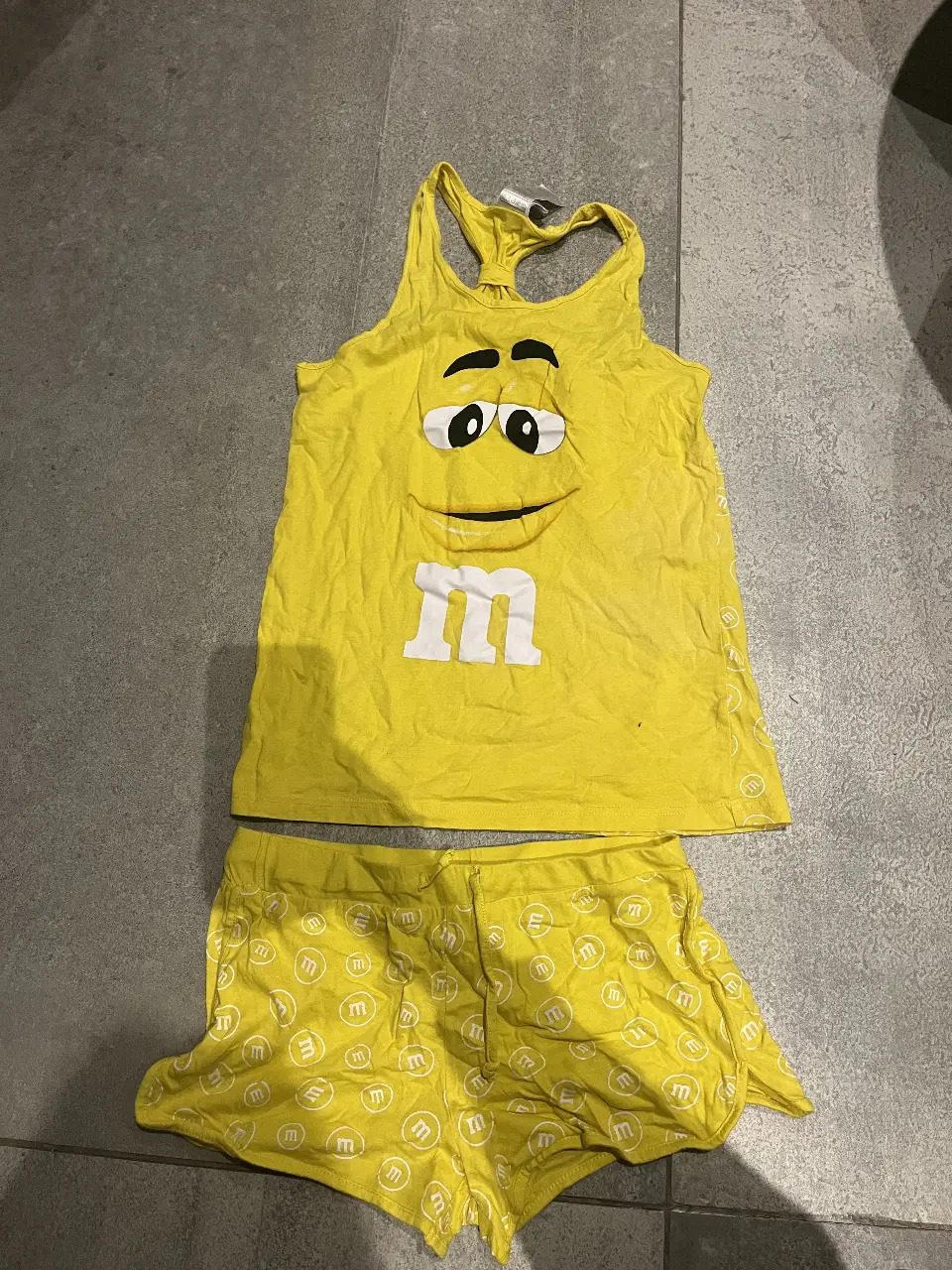 Pijama de dos piezas de M&M's, con musculosa amarilla con estampado de la cara del personaje y shorts a juego con estampado repetido del logo. Ideal para dormir cómodo y con estilo.